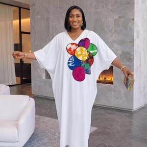 Puede incluir: Vestido blanco con cuello en V y un diseño abstracto colorido en la parte delantera. El vestido tiene mangas cortas y un corte holgado. La mujer lleva sandalias doradas y sostiene un pequeño bolso de mano estampado.