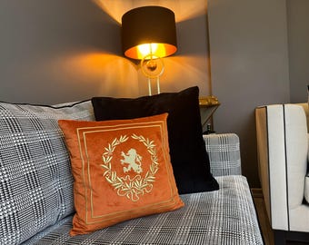 May include: A black and white patterned couch with a black velvet pillow and an orange velvet pillow with an embroidered gold lion design. A gold table lamp with a black shade is in the background.
