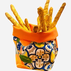 Op de afbeelding: Een fel oranje stoffen mand met een kleurrijk bloemenpatroon met sinaasappels en bladeren. De mand is gevuld met dunne, gouden stokbroodjes bedekt met sesamzaadjes.
