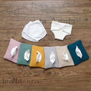 Könnte beinhalten: Eine Auswahl an Babyartikeln, darunter eine weiße Stoffwindel, mehrere weiße Waschlappen und eine Reihe farbiger, quadratischer Waffelstofftücher in Rosa, Grün, Gelb, Grau, Beige und Türkis. Jedes Tuch hat ein weißes, blütenblattförmiges Element.
