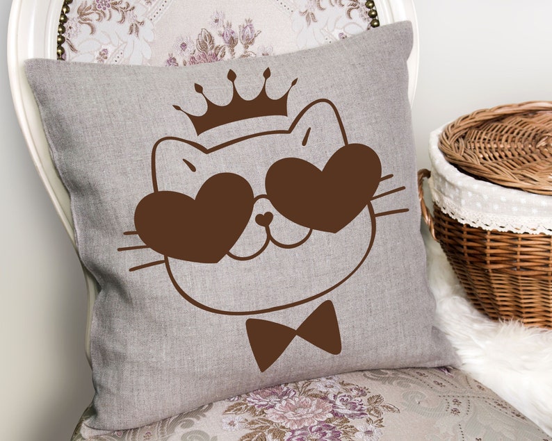 May include: A brown linen pillow with a cute cartoon cat wearing a crown and heart-shaped sunglasses. The cat is wearing a bow tie.