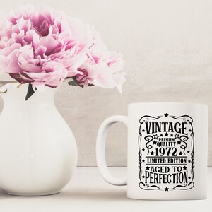 May include: A white ceramic mug with a black graphic that reads "VINTAGE PREMIUM QUALITY 1972 LIMITED EDITION AGED TO PERFECTION." A white vase with pink flowers is next to the mug.