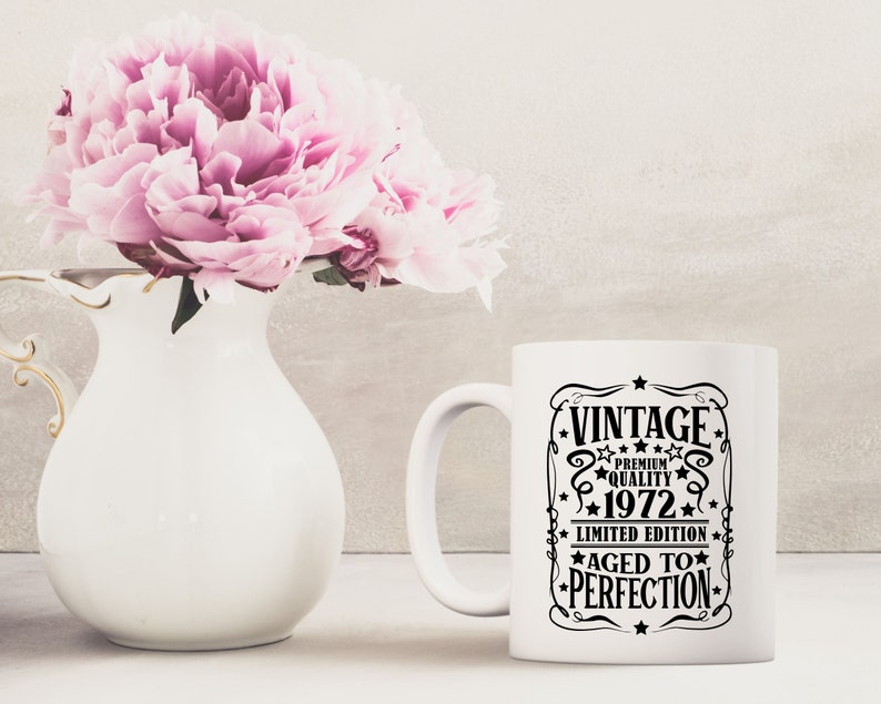 May include: A white ceramic mug with a black graphic that reads "VINTAGE PREMIUM QUALITY 1972 LIMITED EDITION AGED TO PERFECTION." A white vase with pink flowers is next to the mug.