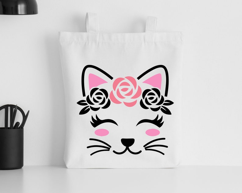 May include: White tote bag with a black and pink silhouette of a cat wearing a flower crown. The cat has closed eyes and a smiling mouth.