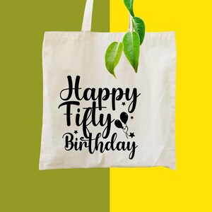 May include: A cream-colored tote bag with the words "Happy Fifty Birthday" in black script. The design includes balloon and star graphics. The bag is hanging with a green and yellow background.