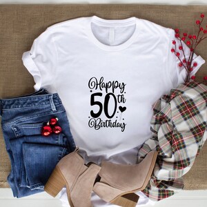 May include: White t-shirt with black text that says "Happy 50th Birthday". The shirt is laying on a brown burlap surface with blue jeans, a plaid scarf, and brown boots.