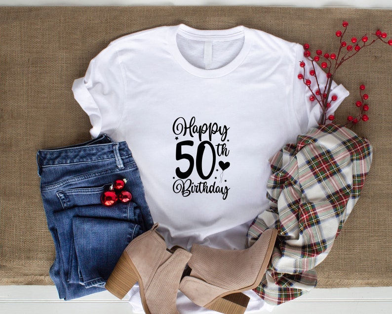 May include: White t-shirt with black text that says "Happy 50th Birthday". The shirt is laying on a brown burlap surface with blue jeans, a plaid scarf, and brown boots.