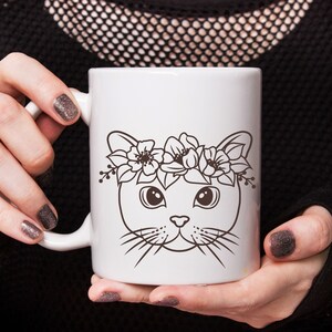 May include: White ceramic mug with a black and white illustration of a cat wearing a flower crown. The cat has large eyes and a cute expression.