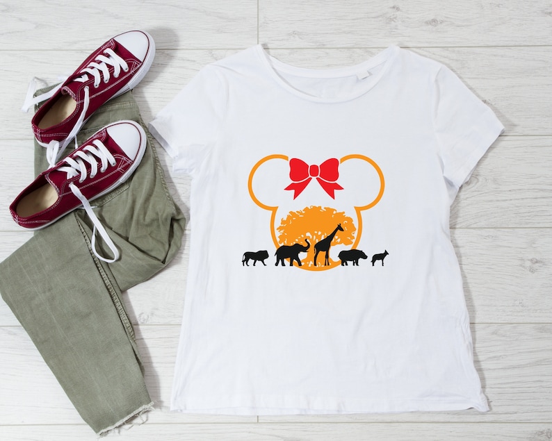 May include: White t-shirt with a yellow Mickey Mouse head silhouette. Inside the head is a brown tree with a red bow on top. There are black silhouettes of animals including a giraffe, elephant, hippopotamus, and a lion.