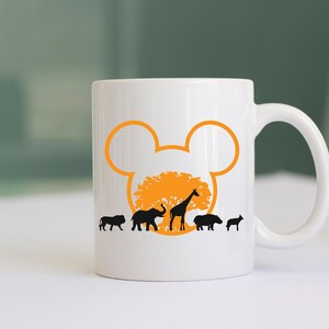 May include: White ceramic coffee mug with a black silhouette of a lion, elephant, giraffe, hippopotamus, and rhinoceros walking in front of a tree with an orange Mickey Mouse ear silhouette behind it.