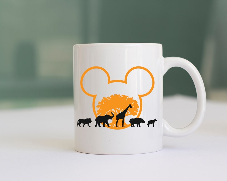 May include: White ceramic coffee mug with a black silhouette of a lion, elephant, giraffe, hippopotamus, and rhinoceros walking in front of a tree with an orange Mickey Mouse ear silhouette behind it.