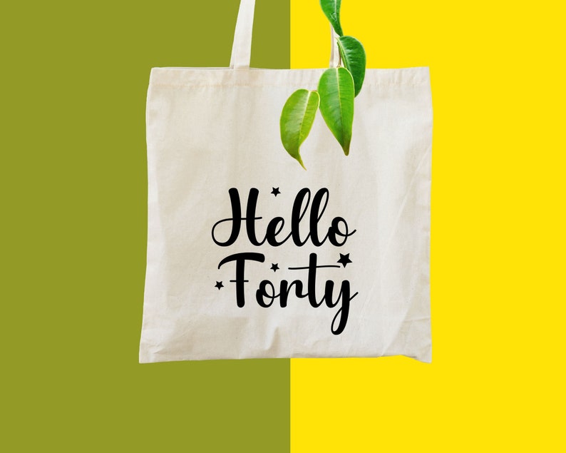 May include: White tote bag with black text that says "Hello Forty" with stars on either side of the text. The bag is hanging from a green plant with leaves.
