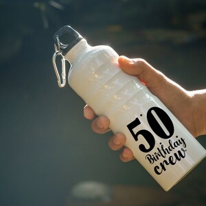 May include: White water bottle with a black lid and a silver carabiner clip. The bottle has the text "50 Birthday Crew" in black. The bottle is held in a person's hand.