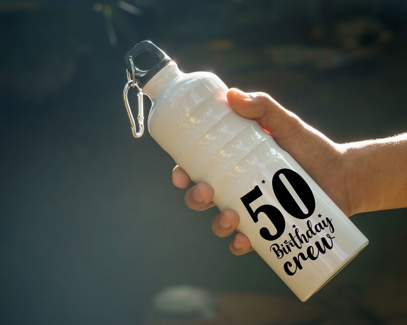 May include: White water bottle with a black lid and a silver carabiner clip. The bottle has the text "50 Birthday Crew" in black. The bottle is held in a person's hand.