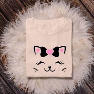 May include: A cream-colored t-shirt with a black and pink cat face graphic. The cat has a pink heart-shaped bow on its head and black eyelashes.