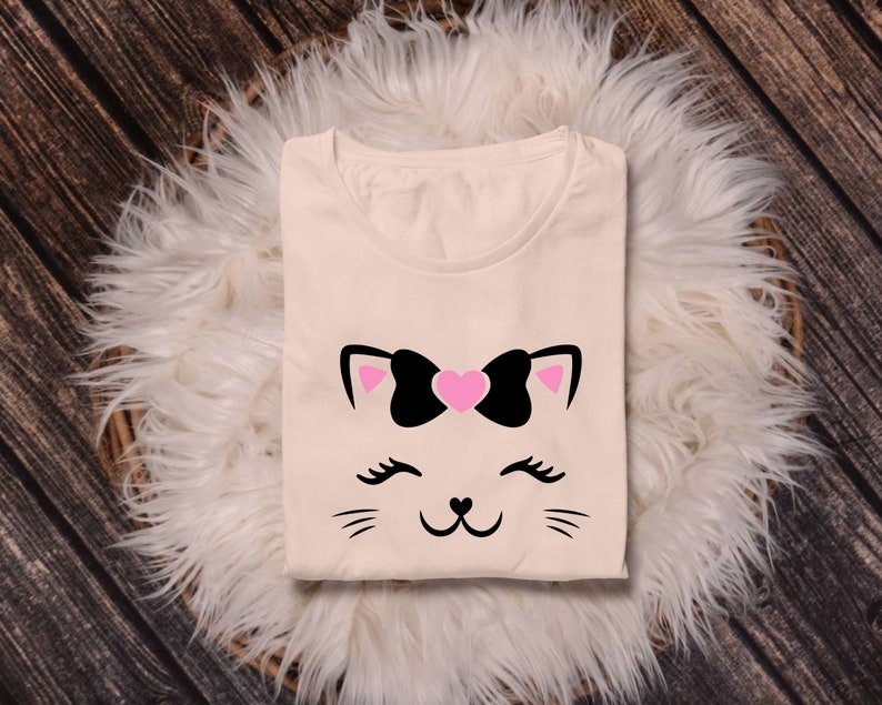 May include: A cream-colored t-shirt with a black and pink cat face graphic. The cat has a pink heart-shaped bow on its head and black eyelashes.
