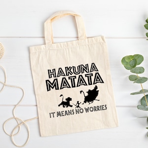 May include: A white canvas tote bag with black text and silhouettes of characters from the movie The Lion King. The text reads "Hakuna Matata" and "It means no worries".