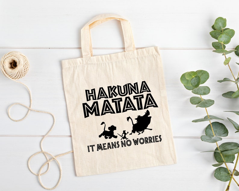 May include: A white canvas tote bag with black text and silhouettes of characters from the movie The Lion King. The text reads "Hakuna Matata" and "It means no worries".