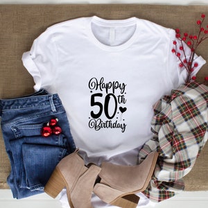 May include: White t-shirt with the text "Happy 50th Birthday" in black script. The shirt is paired with blue jeans, beige ankle boots, and a plaid scarf. Red Christmas ornaments are in the jeans pocket.