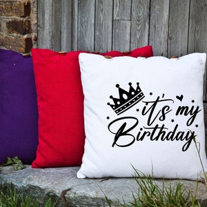 K&ouml;nnte beinhalten: Drei dekorative Kissen mit Knopf-Akzenten. Das wei&szlig;e Kissen hat eine schwarze Krone und den Text "It's my Birthday". Die anderen Kissen sind rot und lila.