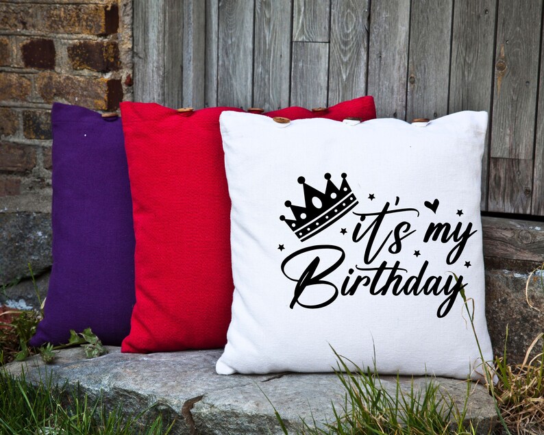 K&ouml;nnte beinhalten: Drei dekorative Kissen mit Knopf-Akzenten. Das wei&szlig;e Kissen hat eine schwarze Krone und den Text "It's my Birthday". Die anderen Kissen sind rot und lila.