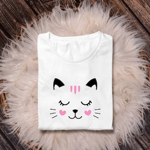 May include: White t-shirt with a black and pink cat face graphic. The cat has closed eyes and pink hearts on its cheeks.