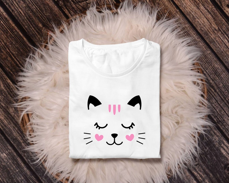 May include: White t-shirt with a black and pink cat face graphic. The cat has closed eyes and pink hearts on its cheeks.