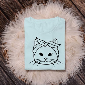 May include: A light blue t-shirt with a black and white illustration of a cat wearing a headband. The cat has large eyes and a cute expression.