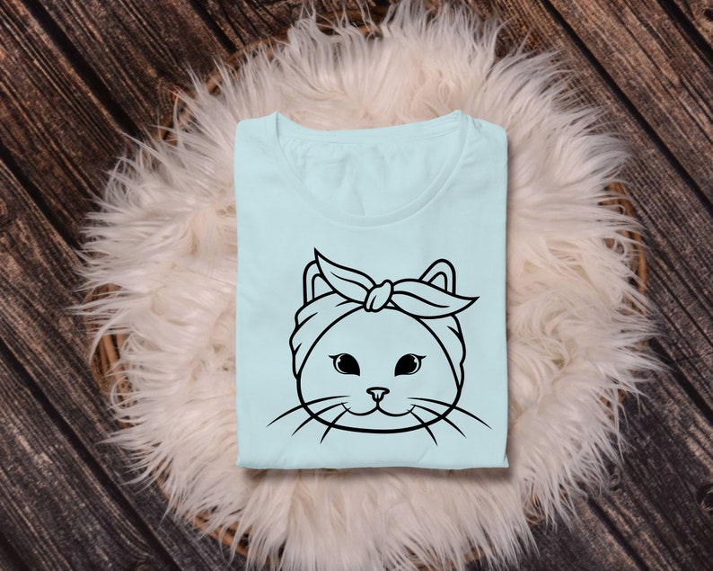 May include: A light blue t-shirt with a black and white illustration of a cat wearing a headband. The cat has large eyes and a cute expression.
