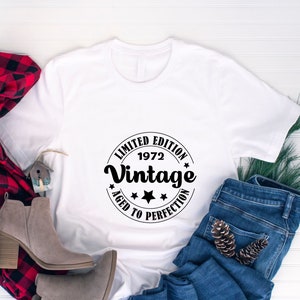 May include: White t-shirt with a black circular graphic that reads "LIMITED EDITION 1972 Vintage AGED TO PERFECTION". The shirt is paired with blue jeans, a red and black plaid scarf, and gray ankle boots.
