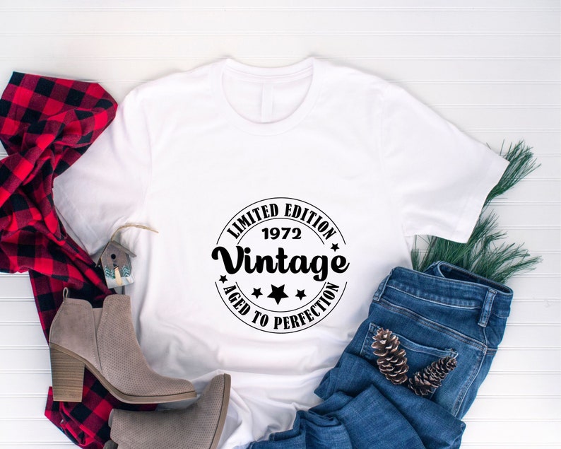 May include: White t-shirt with a black circular graphic that reads "LIMITED EDITION 1972 Vintage AGED TO PERFECTION". The shirt is paired with blue jeans, a red and black plaid scarf, and gray ankle boots.