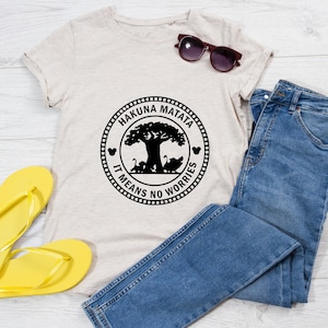 May include: A light beige t-shirt with a black circle graphic featuring the words "Hakuna Matata" and "It Means No Worries" around a silhouette of a tree and animals. The t-shirt is laid out on a white wooden surface with a pair of yellow flip-flops and blue jeans.