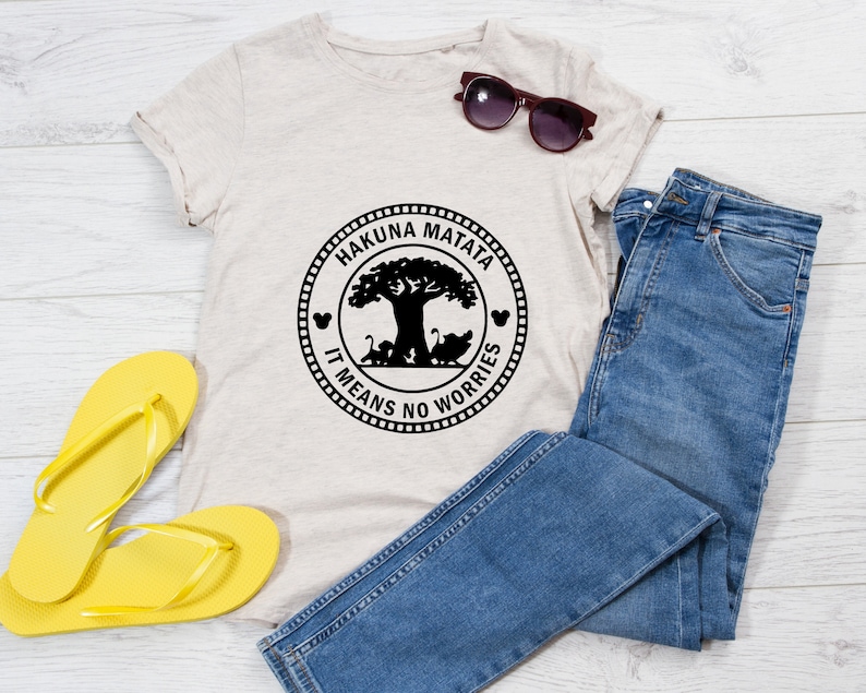 May include: A light beige t-shirt with a black circle graphic featuring the words "Hakuna Matata" and "It Means No Worries" around a silhouette of a tree and animals. The t-shirt is laid out on a white wooden surface with a pair of yellow flip-flops and blue jeans.
