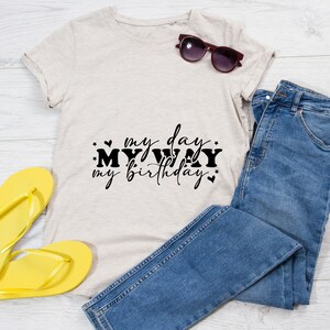 K&ouml;nnte beinhalten: Ein beiges T-Shirt mit dem Text "my day my way my birthday" in schwarzer Schrift. Das Shirt liegt auf einer wei&szlig;en Holzoberfl&auml;che mit einem Paar gelber Flip-Flops und einer blauen Jeans.