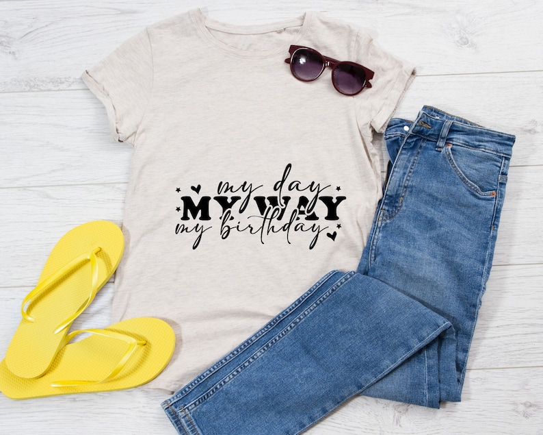 K&ouml;nnte beinhalten: Ein beiges T-Shirt mit dem Text "my day my way my birthday" in schwarzer Schrift. Das Shirt liegt auf einer wei&szlig;en Holzoberfl&auml;che mit einem Paar gelber Flip-Flops und einer blauen Jeans.