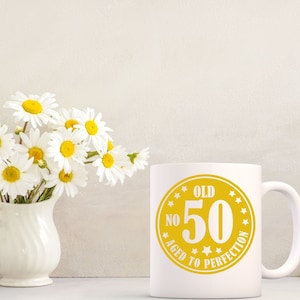 May include: White ceramic mug with a yellow graphic that reads "OLD NO 50 AGED TO PERFECTION". A bouquet of white daisies with yellow centers sits in a white vase. The mug is on a white surface.