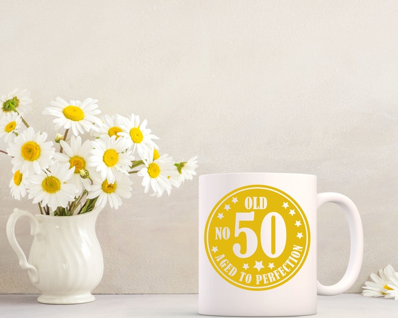 May include: White ceramic mug with a yellow graphic that reads "OLD NO 50 AGED TO PERFECTION". A bouquet of white daisies with yellow centers sits in a white vase. The mug is on a white surface.