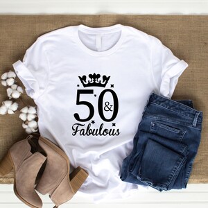 May include: White t-shirt with the text "50 & Fabulous" in black script, adorned with a crown and stars. The shirt is paired with blue jeans and tan ankle boots. The items are arranged on a brown burlap surface.