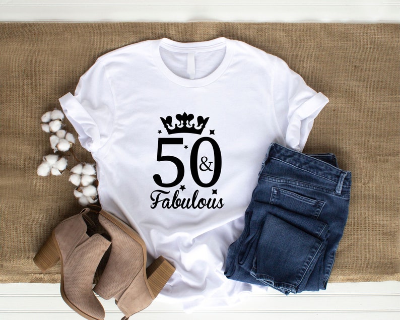 May include: White t-shirt with the text "50 & Fabulous" in black script, adorned with a crown and stars. The shirt is paired with blue jeans and tan ankle boots. The items are arranged on a brown burlap surface.