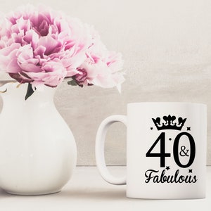 May include: White ceramic mug with a black design that says "40 & Fabulous" with a crown above the number 40. The mug is sitting on a table with a white vase with pink flowers in the background.