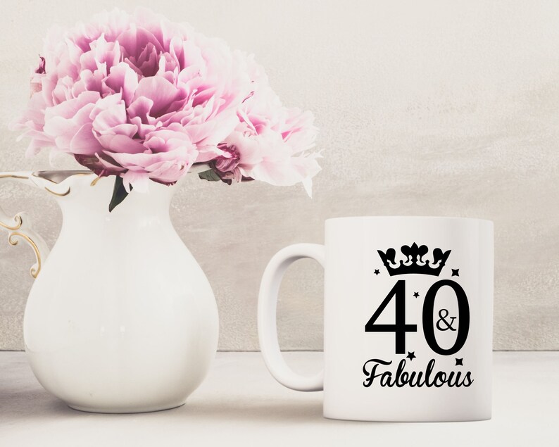 May include: White ceramic mug with a black design that says "40 & Fabulous" with a crown above the number 40. The mug is sitting on a table with a white vase with pink flowers in the background.