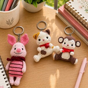 May include: Three crochet keychains featuring a pink piglet with a dark red scarf, a white and brown calico cat with a red collar and bell, and a brown and white monkey with a red scarf, each attached to a silver metal split ring keychain.
