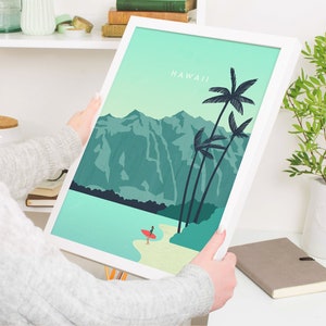 May include: A white framed print with a teal and green landscape scene of mountains, palm trees, and a beach with a person surfing. The text "HAWAII" is at the top of the print.