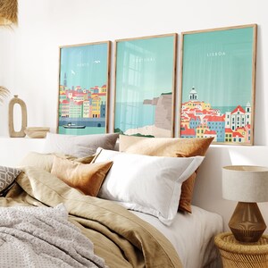 May include: Three framed art prints featuring colorful illustrations of Portuguese cityscapes. Each print has a light blue background with the city name at the top. The prints are displayed above a bed with neutral-toned bedding and pillows.
