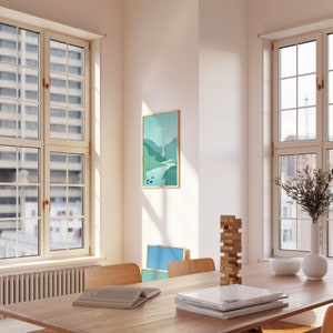 May include: A framed landscape art print with a waterfall scene in shades of green and blue hangs on a white wall. The print is in a room with a wooden table, chairs, and windows. A stack of magazines and a book are on the table.
