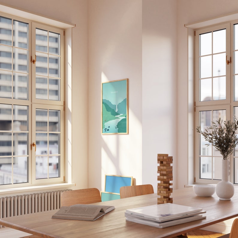 May include: A framed landscape art print with a waterfall scene in shades of green and blue hangs on a white wall. The print is in a room with a wooden table, chairs, and windows. A stack of magazines and a book are on the table.