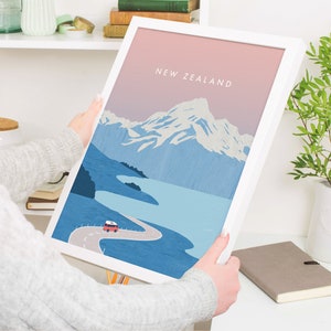 May include: A white framed print with a pink and blue illustration of a mountain range, a lake, and a winding road with a red camper van driving on it. The text "NEW ZEALAND" is printed in black at the top of the print.