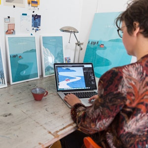 May include: A person working on a laptop computer with a red car on a winding road on the screen. The laptop is on a wooden table with three framed prints of landscapes with the word "Sweden" on one of them.
