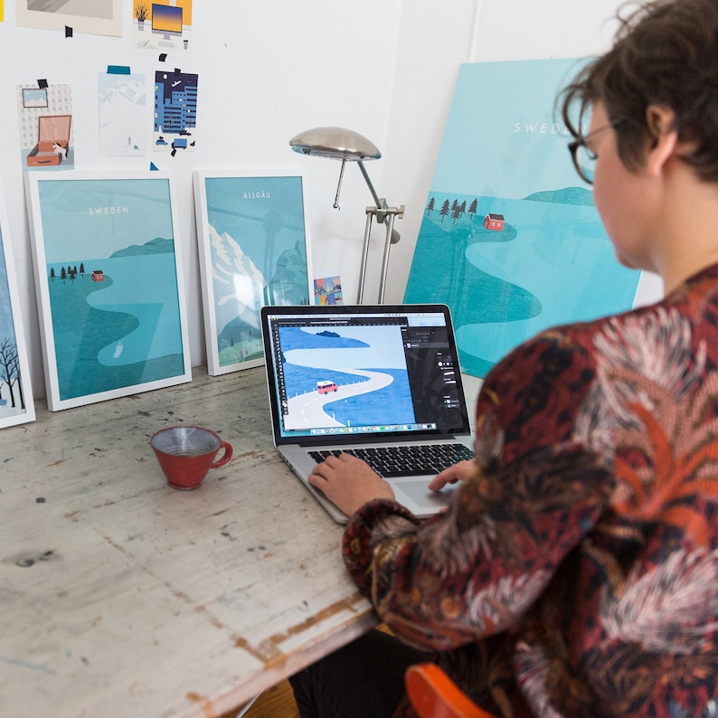 May include: A person working on a laptop computer with a red car on a winding road on the screen. The laptop is on a wooden table with three framed prints of landscapes with the word "Sweden" on one of them.