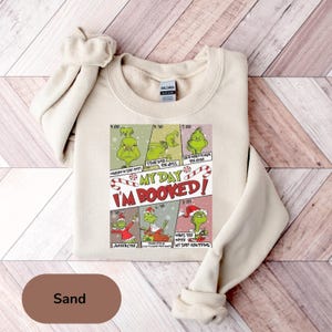 Könnte beinhalten: Sandfarbenes Sweatshirt mit einer Grafik mit dem Grinch und dem Text "MY DAY I'M BOOKED!". Die Grafik enthält verschiedene Grinch-Illustrationen mit Text wie "Wallow in self-pity" und "Stare into the abyss".